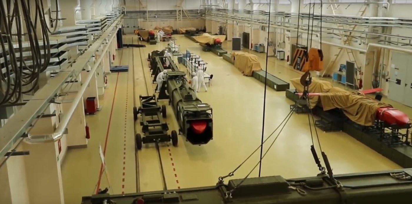 Wide factory hall with workers in white suits, missile sections under tarpaulins and a red nose section on a stand; a video frame of unconfirmed location and date