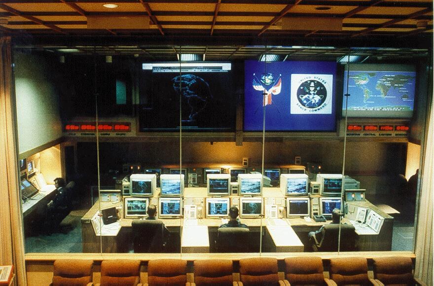 Console room seen through a glass wall with a U.S. Space Command seal on a large screen